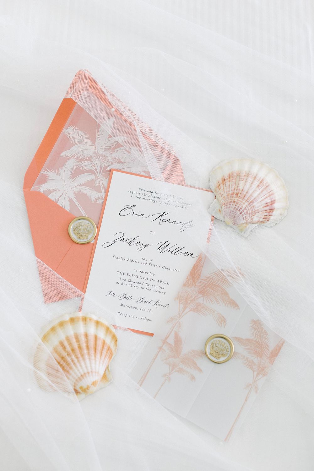 Tropical beach wedding invitation suite with coral palm tree envelope, vellum overlay, gold wax seals, and seashells on white fabric.