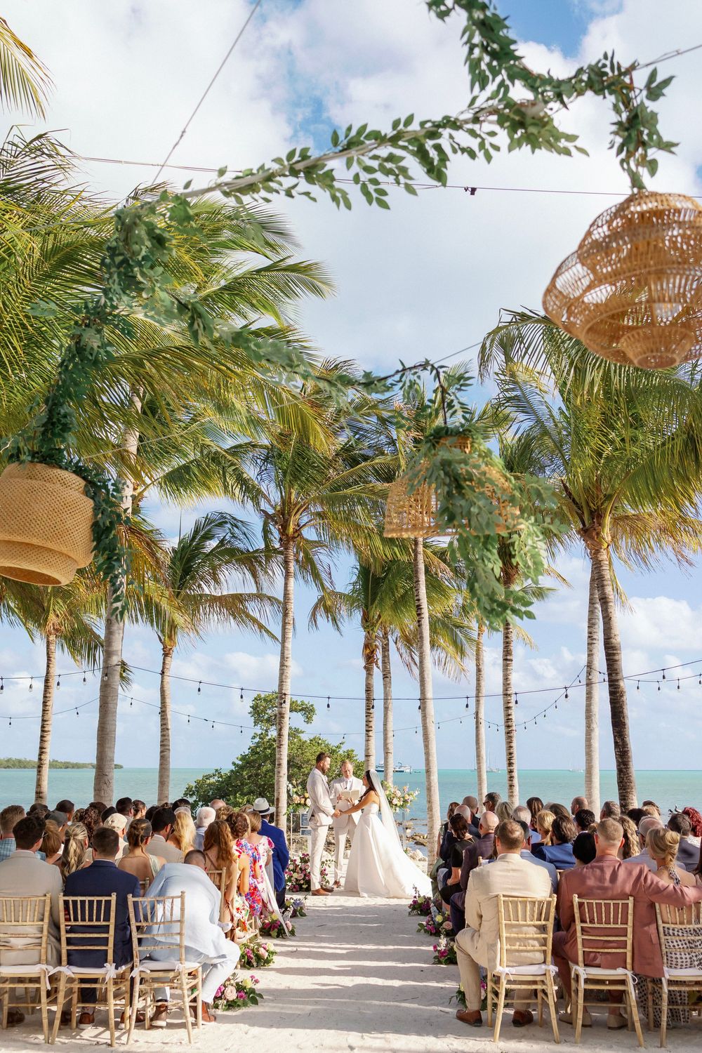 Outdoor beach wedding ceremony with palm trees, string lights, and wicker lanterns overlooking turquoise ocean waters.