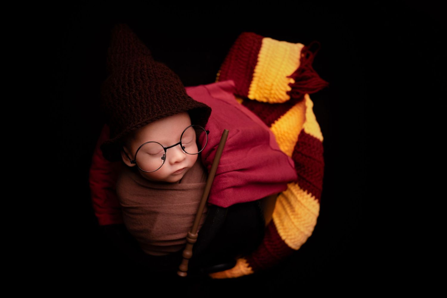 Newborn dressed in Harry Potter themed costume with glasses against dark background.