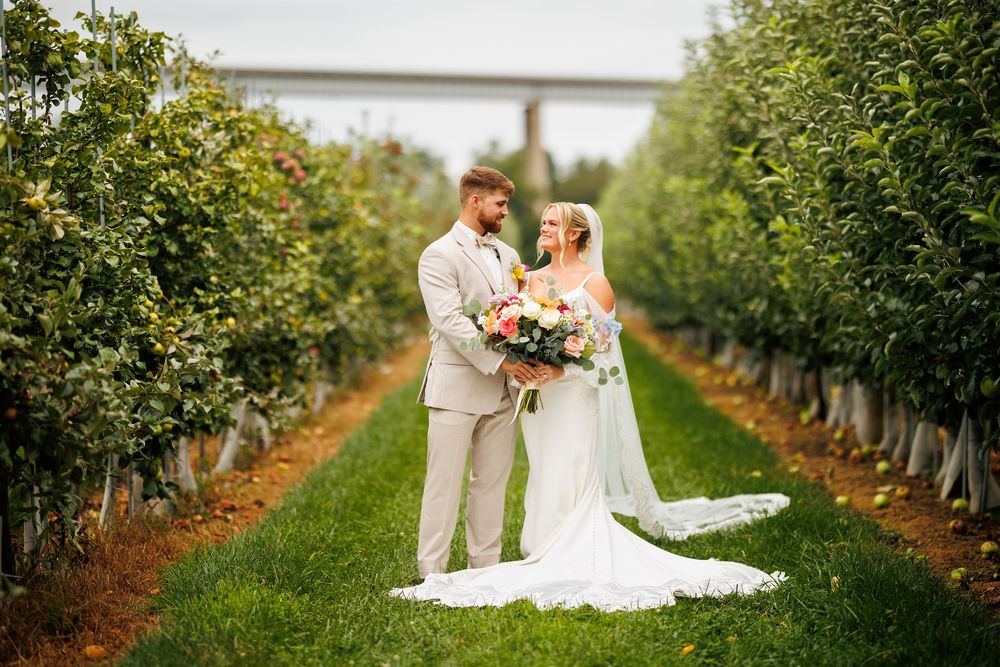 Bride and groom stand together in lush orchard rows, bride holding colorful bouquet in white gown, groom in tan suit.
