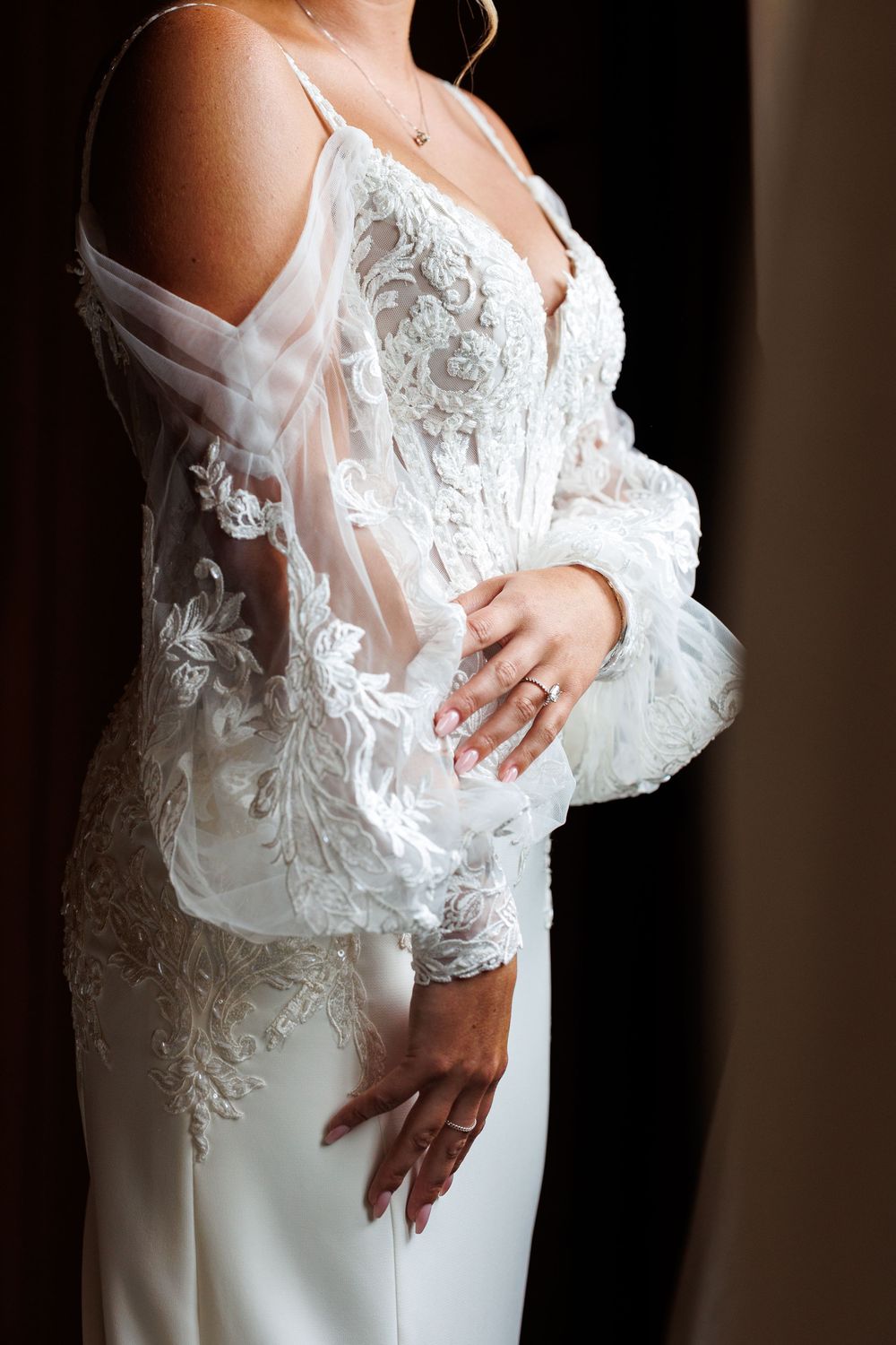 Bride in white lace wedding gown with sheer embroidered sleeves and elegant neckline, wearing a diamond ring.