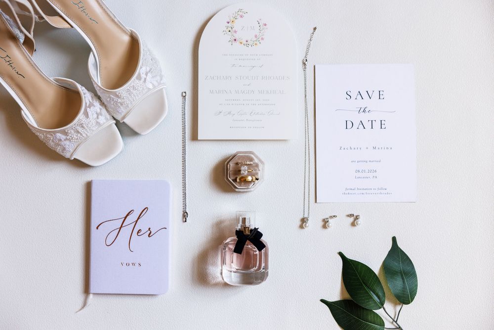 Elegant wedding flat lay with lace heels, save the date card, perfume, jewelry, and floral invitation on white background.