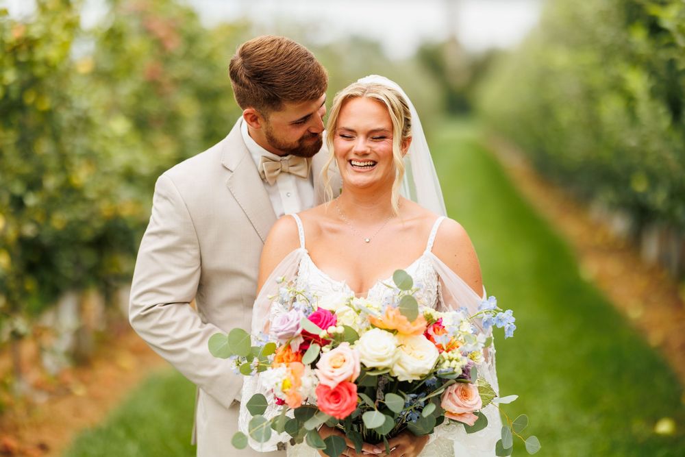 Smiling bride holding colorful bouquet with groom in tan suit embracing her in lush green orchard vineyard.