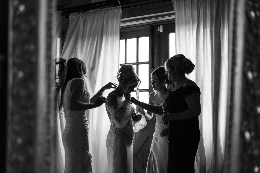 Black and white photo of bride being helped into wedding dress by bridesmaids near a sunlit window.