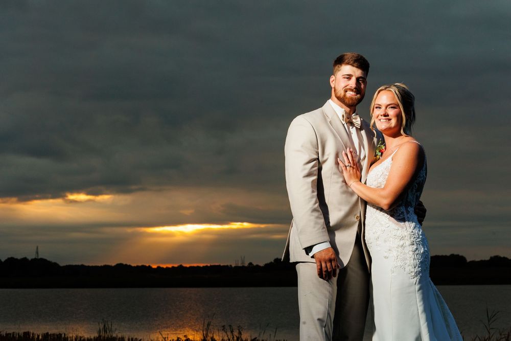 Bride and groom pose romantically by a lake at sunset with dramatic storm clouds in the background.