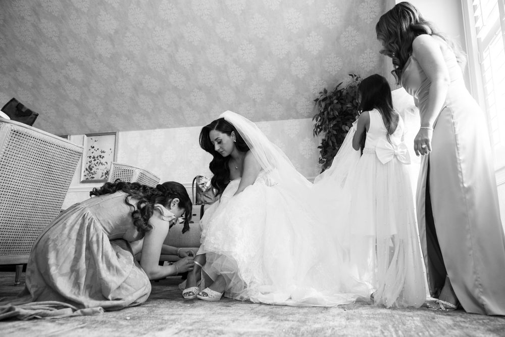 Black and white photo of a bride being helped into her wedding dress by bridesmaids in an elegant room.