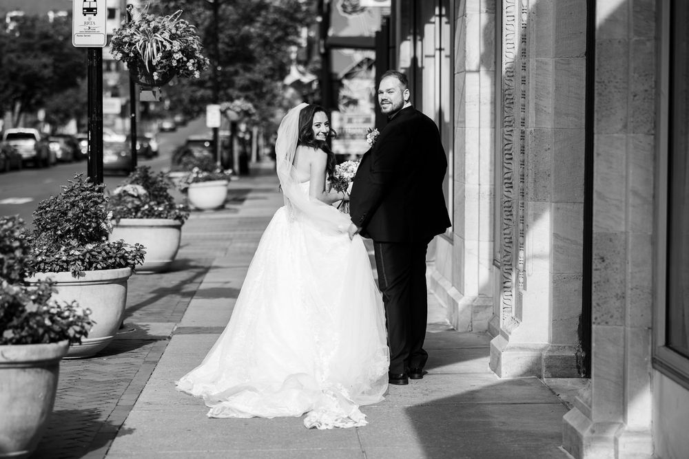 Black and white photo of a bride and groom posing on a city sidewalk, the bride in a flowing gown.