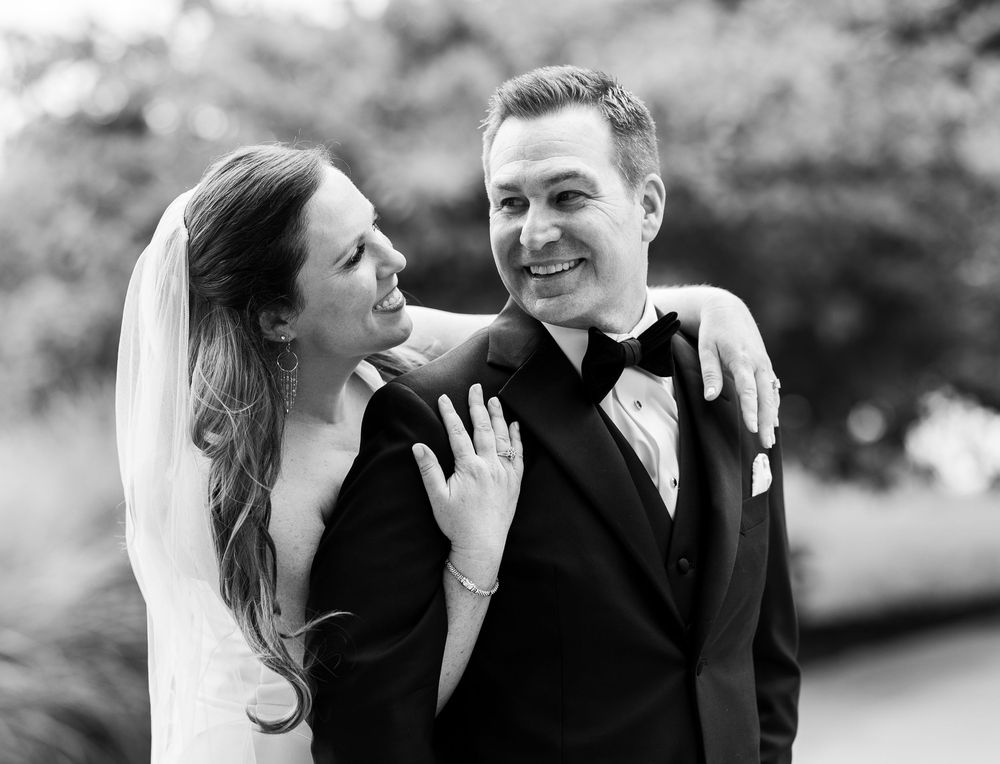 Black and white wedding photo of smiling bride and groom, bride wearing veil and showing ring, groom in tuxedo.