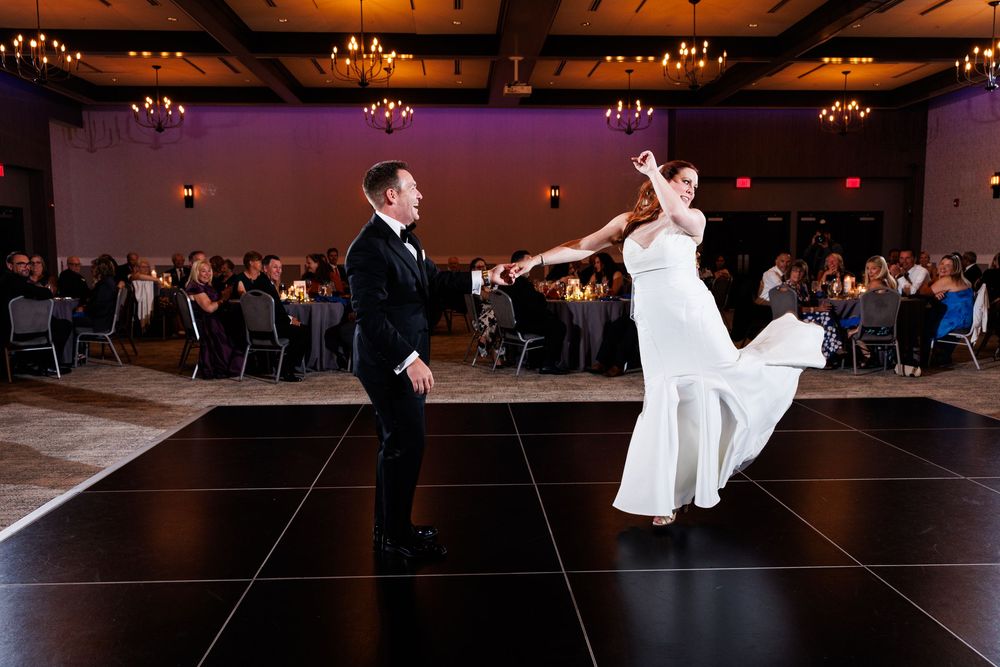Bride and groom share first dance on black dance floor at elegant wedding reception with guests watching.