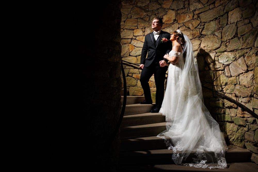 Bride and groom stand on stone staircase in dramatic lighting, bride in lace gown with veil, groom in black tuxedo.