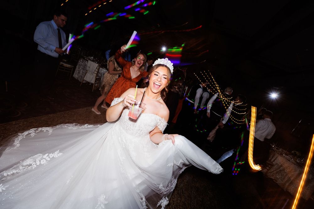 A joyful bride in a white wedding gown dances energetically at a reception, surrounded by guests with colorful glow sticks.