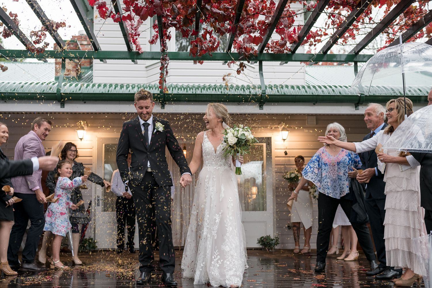 A newlywed couple is walking hand in hand under a pergola adorned with flowers. They are surrounded by guests who are joyfully throwing conf