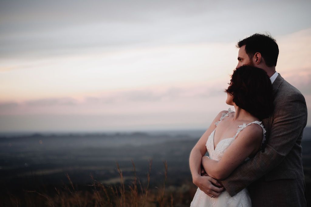 A couple embraces on a hilltop at sunset, silhouetted against a dusky sky over a scenic mountain landscape. Wouter Kleynhans Photography