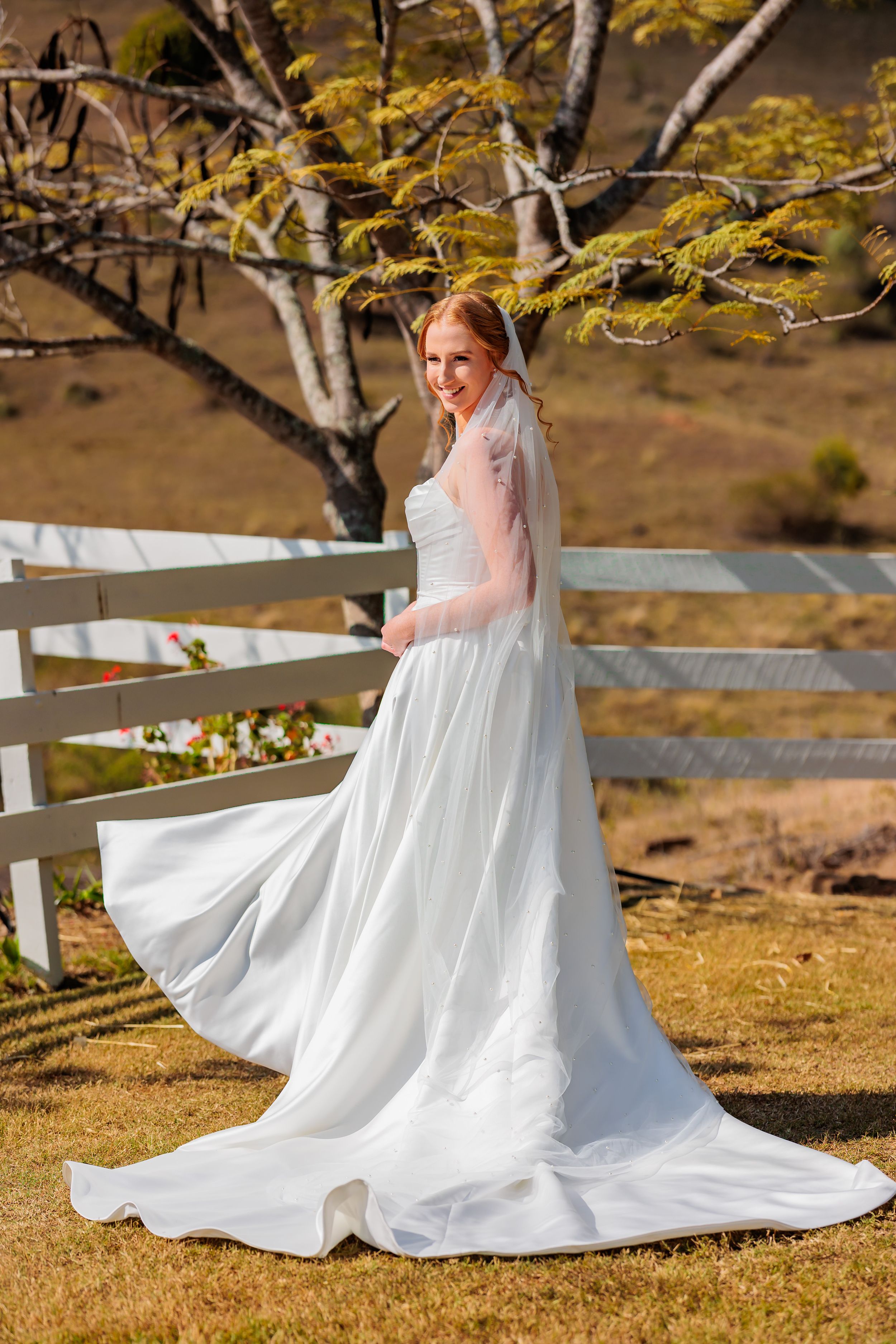 Smiling bride in white gown and veil poses by rustic fence ar Branell Homestead in Laidley Qld Wedding photography