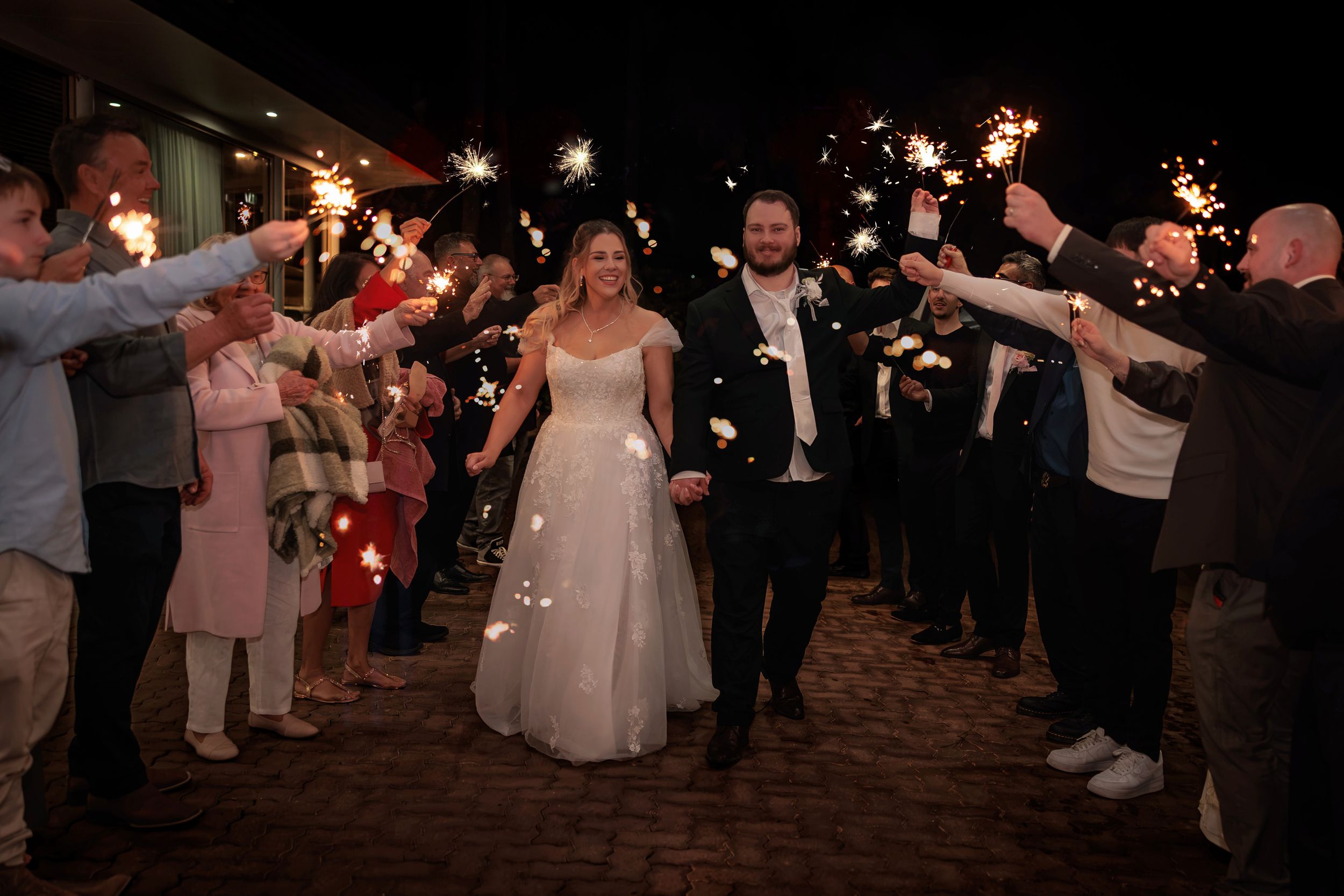 Bride and groom walk through sparkler exit tunnel at night wedding celebration with guests lining the path. Wedding Photography