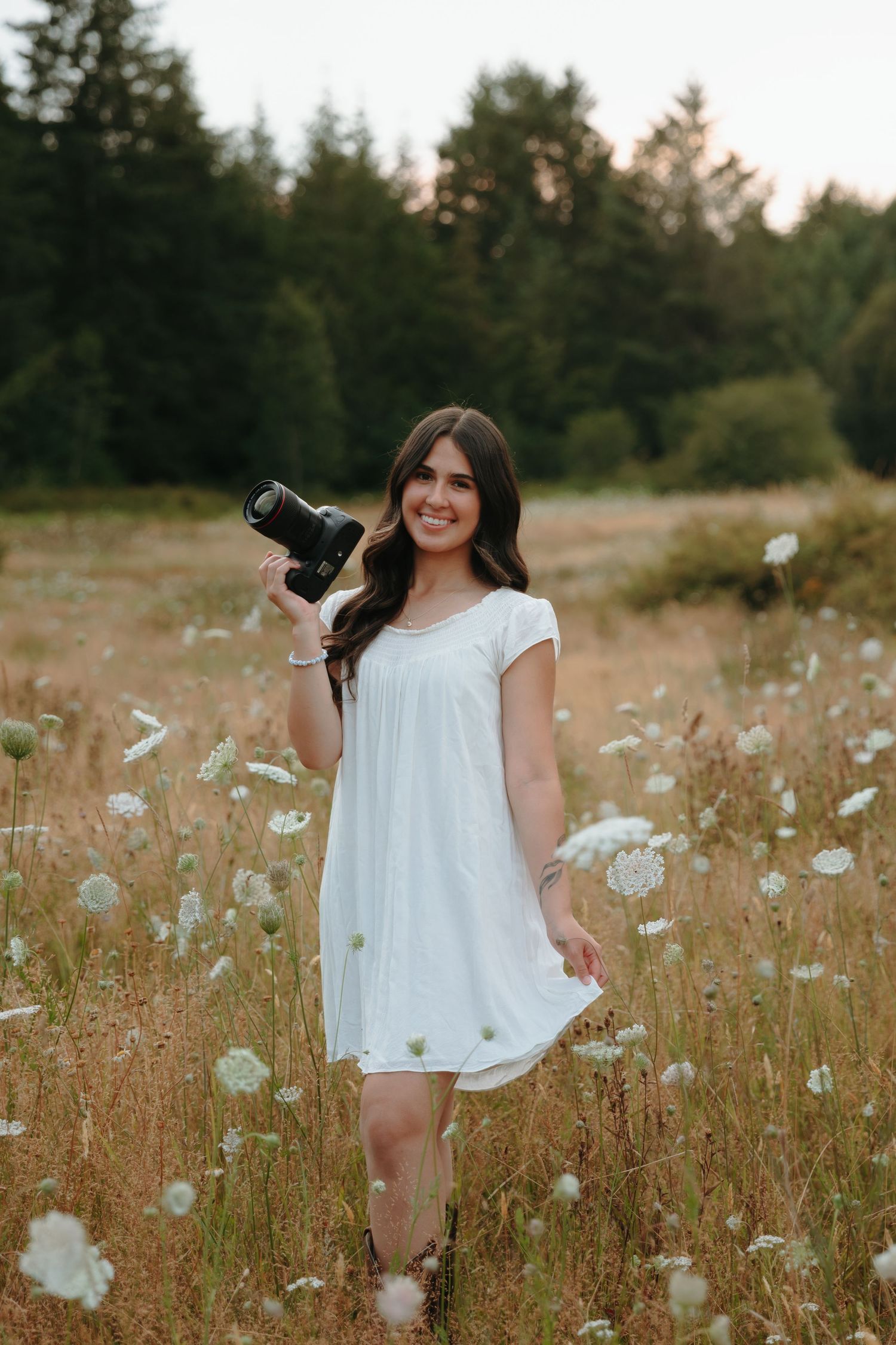 A photographer in a white dress stands in a field of wild white flowers during golden hour.