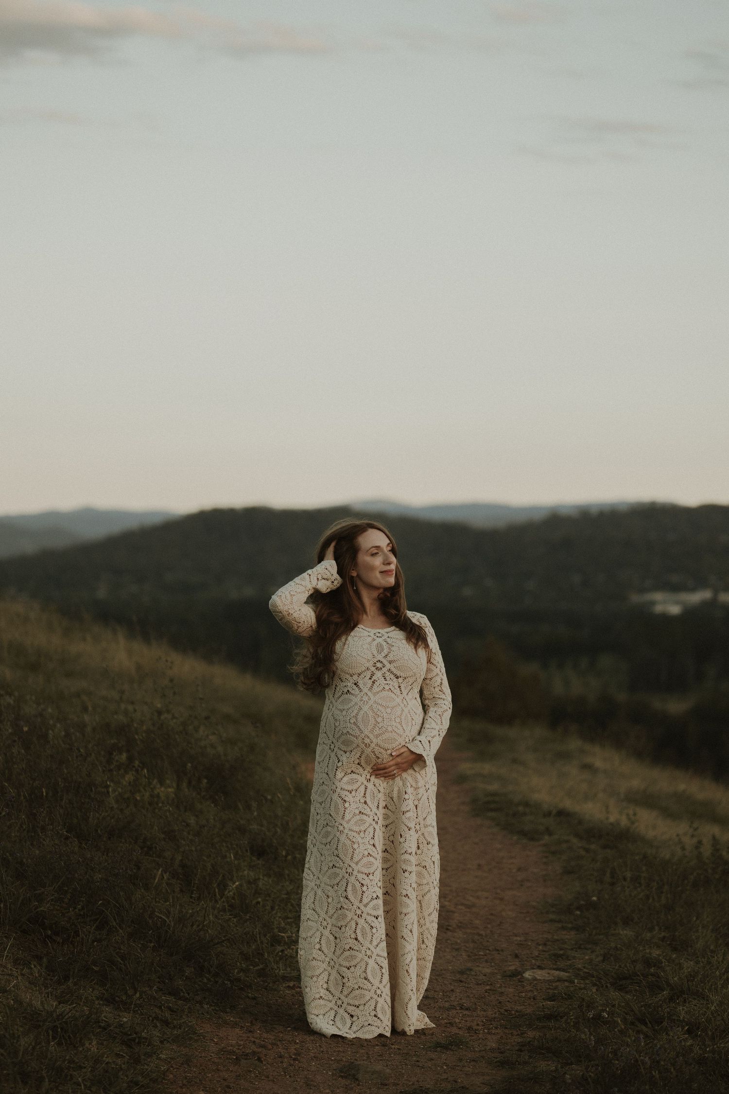 Expectant mother in flowing white lace dress stands on hilltop trail at dusk against mountain landscape.