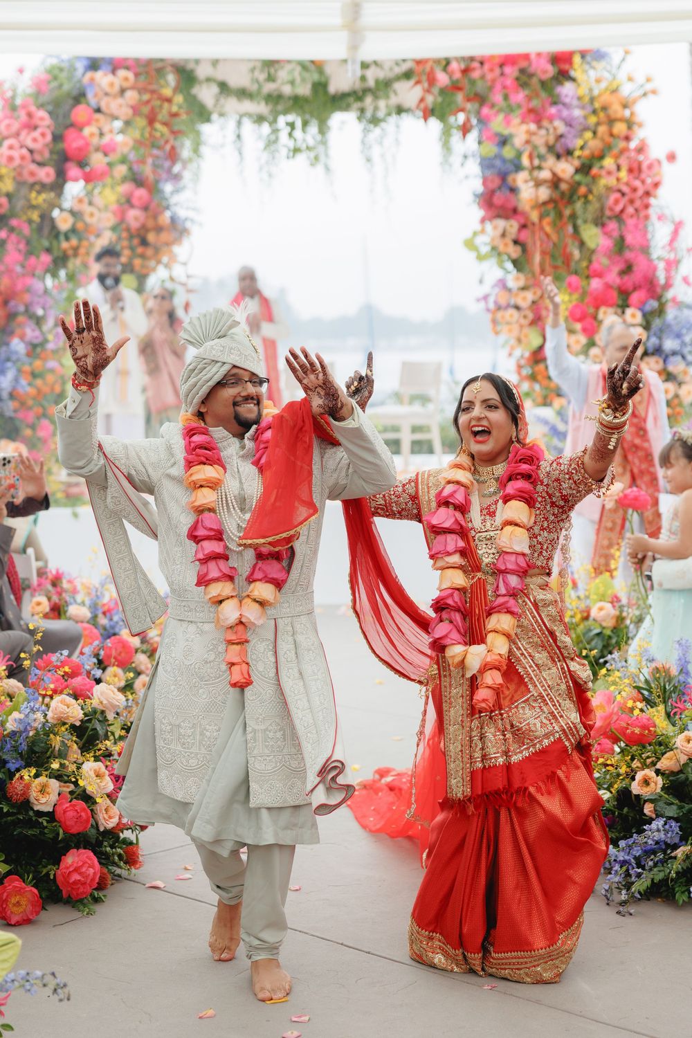 A joyful couple in traditional wedding attire celebrates during their ceremony beneath pink floral decorations.
