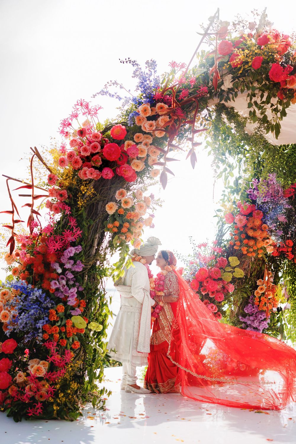 A wedding photoshoot series featuring flowing red fabric against a lush floral arch with pink, purple and red blooms.