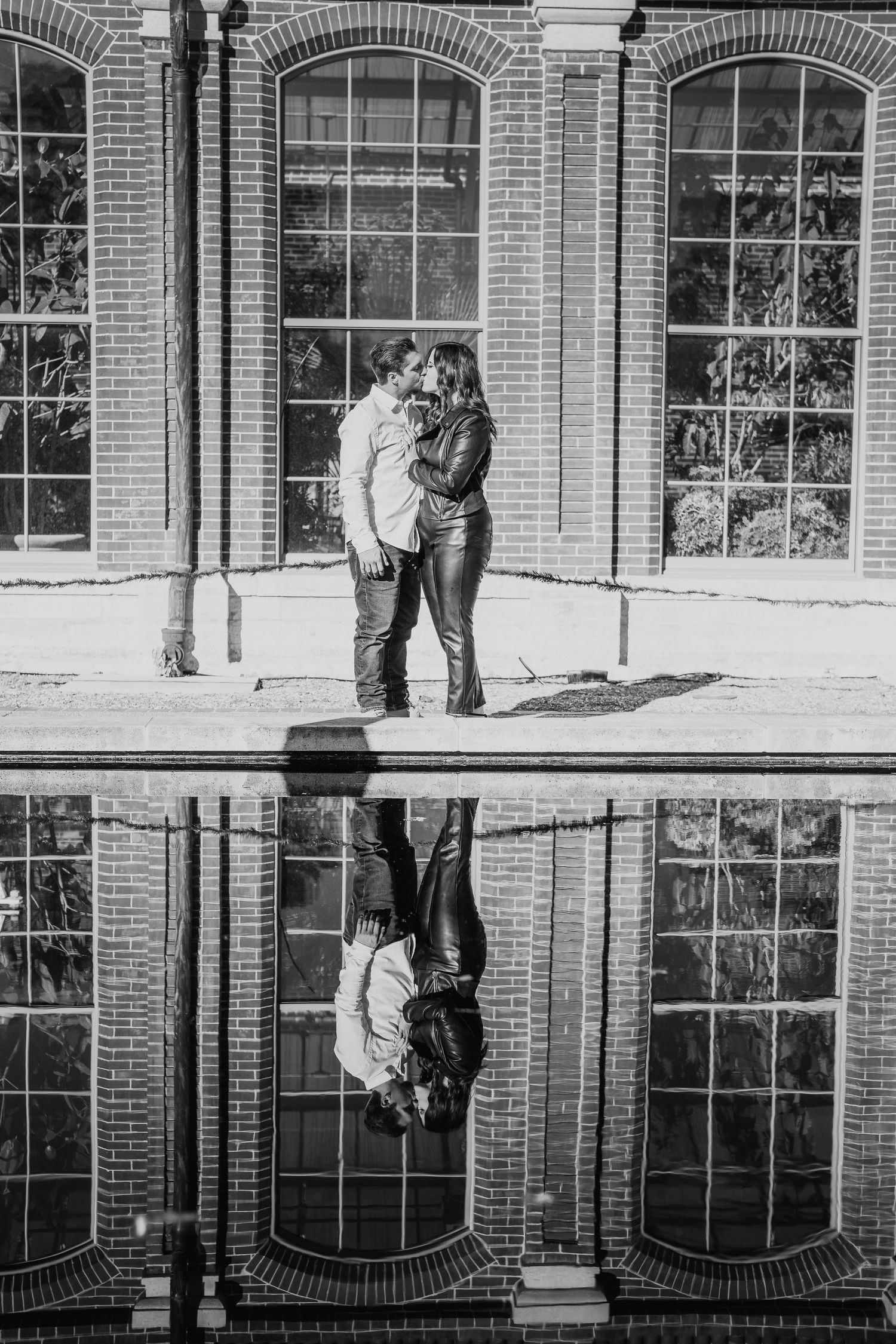 A couple's reflection appears in a puddle beneath brick architecture with arched windows.