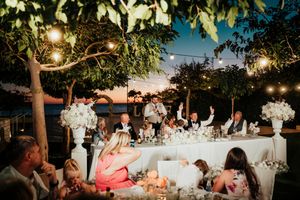 String lights illuminate an elegant outdoor wedding reception setup with white tablecloths and floral centerpieces at dusk.