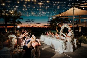 Wedding reception celebration at sunset under string lights on an outdoor terrace with palm trees and ocean views.