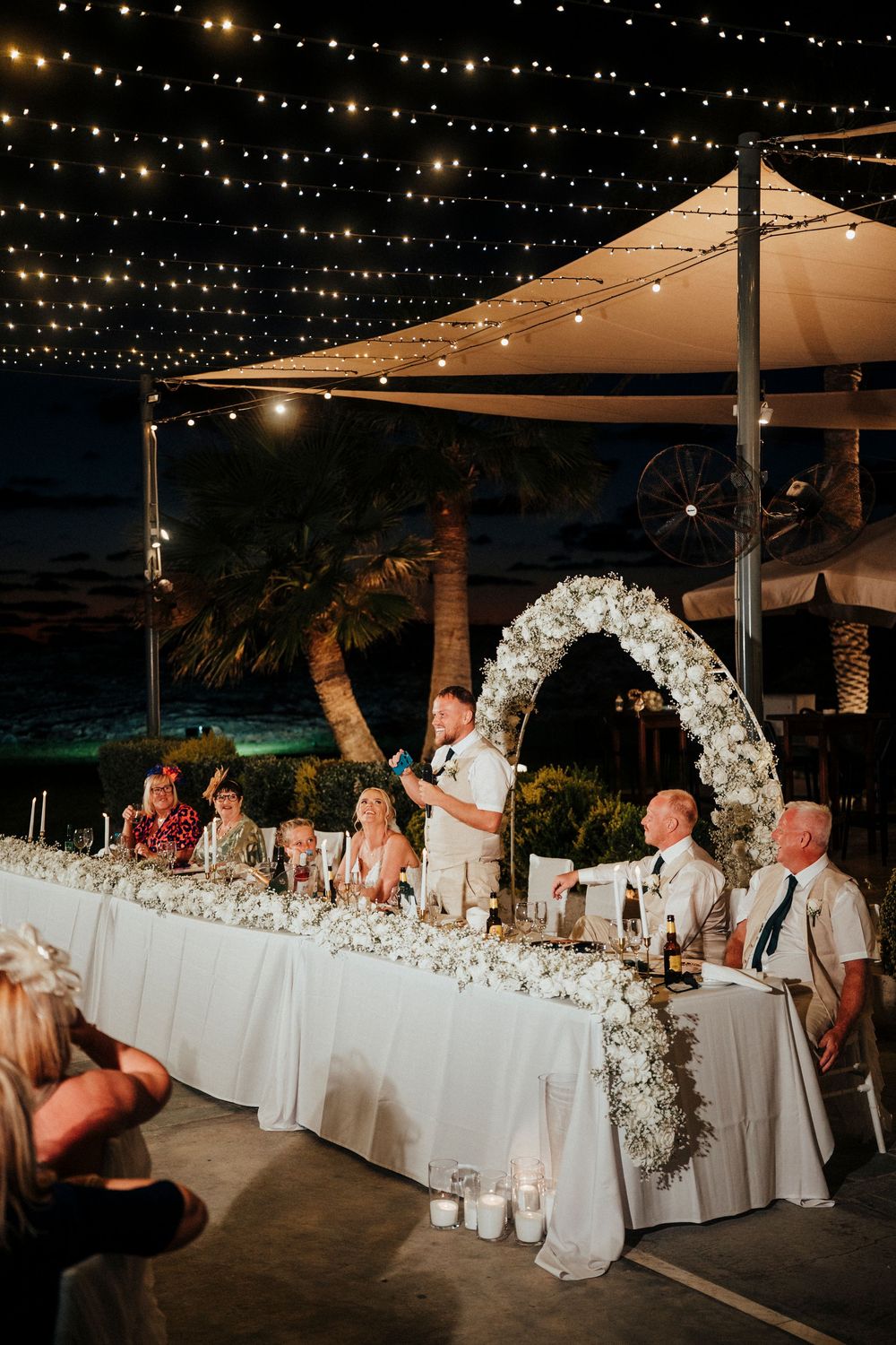 Wedding reception setup at night with string lights overhead and a long white banquet table decorated with floral arrangements.