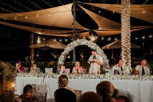 Wedding reception at night with illuminated floral arch and string lights over head table at outdoor venue.
