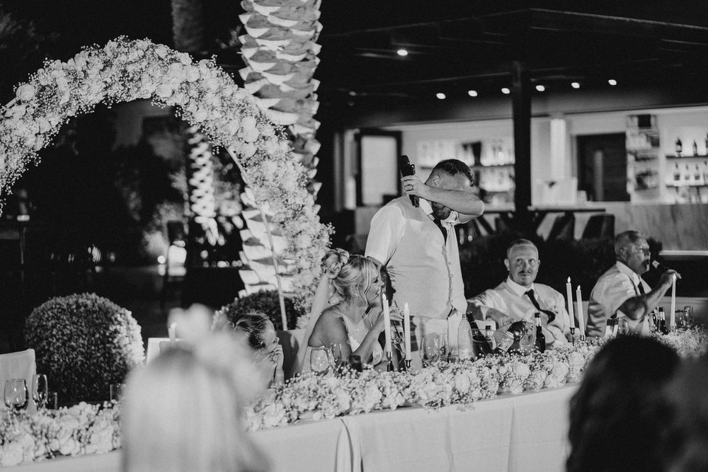 Black and white photo of a wedding reception with an elegant floral arch and head table decorated with flowers.