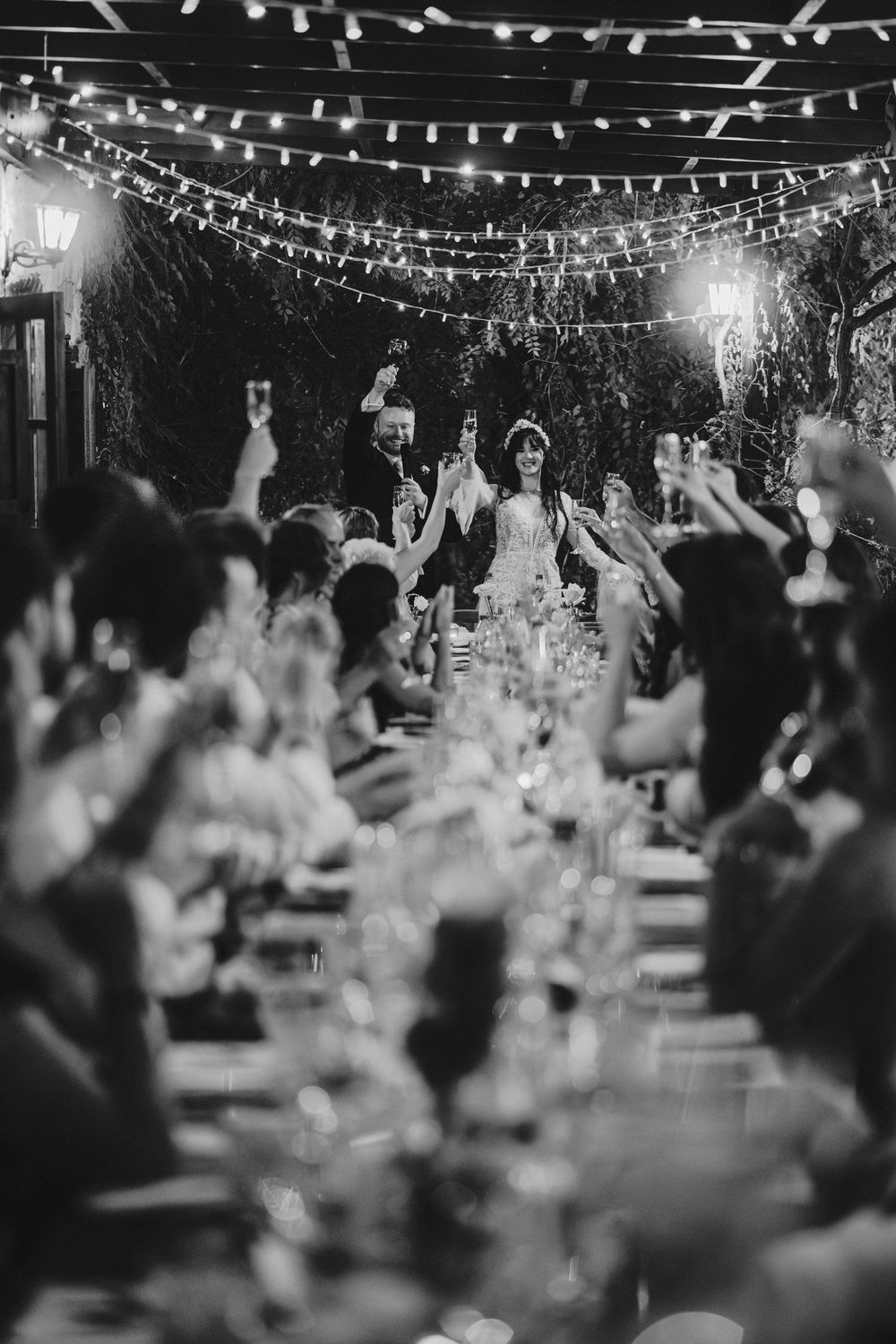 String lights illuminate an outdoor wedding reception as guests raise their hands in celebration at dusk.