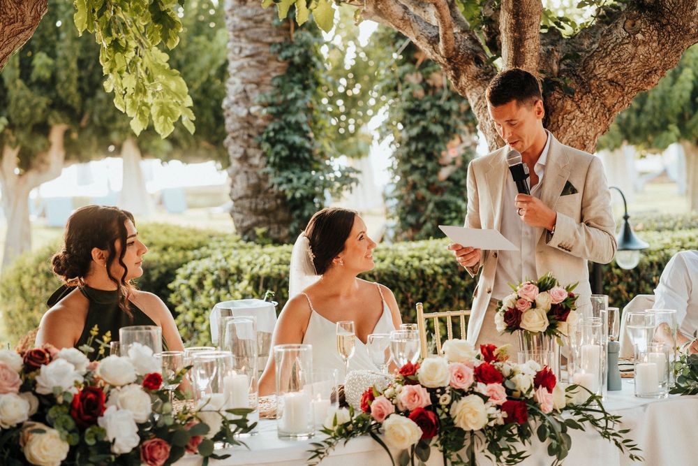 An outdoor wedding reception under palm trees with elegant table settings and pink and white floral centerpieces.