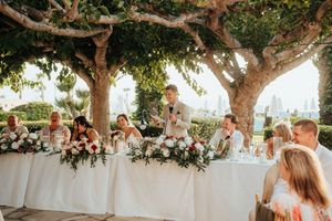 Wedding guests dine at elegant outdoor reception tables under a canopy of trees with string lights at sunset.