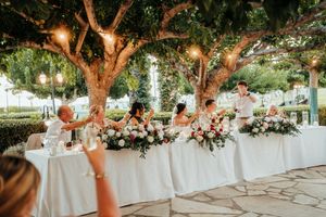 Outdoor wedding reception setup with string lights in trees and elegant floral arrangements on white-clothed tables at dusk.