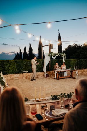 String lights illuminate an outdoor wedding reception venue at dusk with cypress trees in the background.