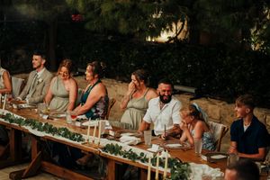 Wedding guests seated at a long outdoor table decorated with greenery and candles during a sunset dinner reception.