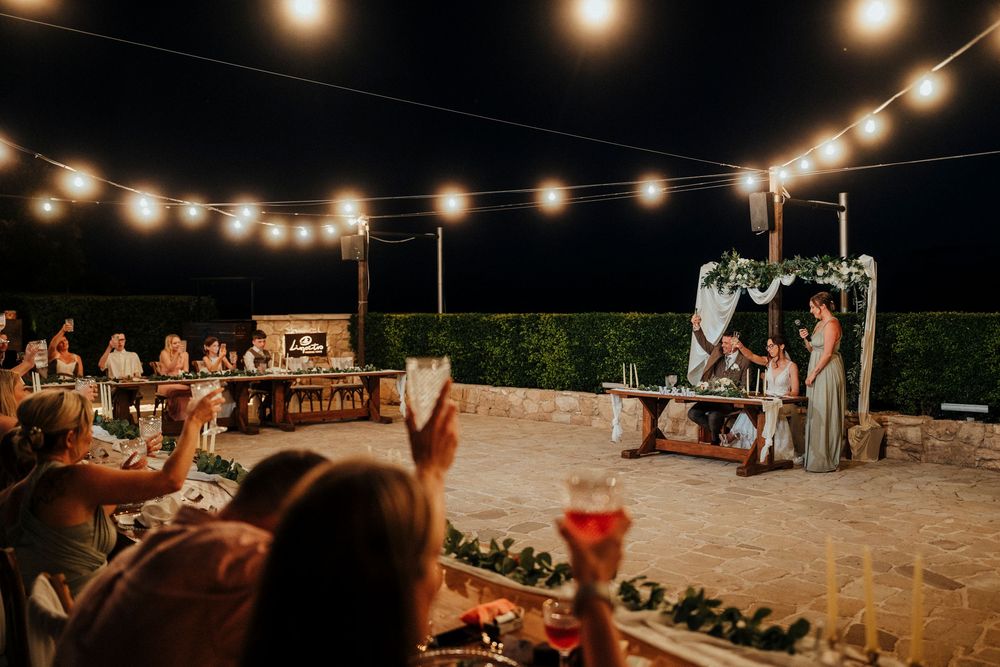 Nighttime outdoor celebration with glowing string lights and guests gathered at long wooden tables.