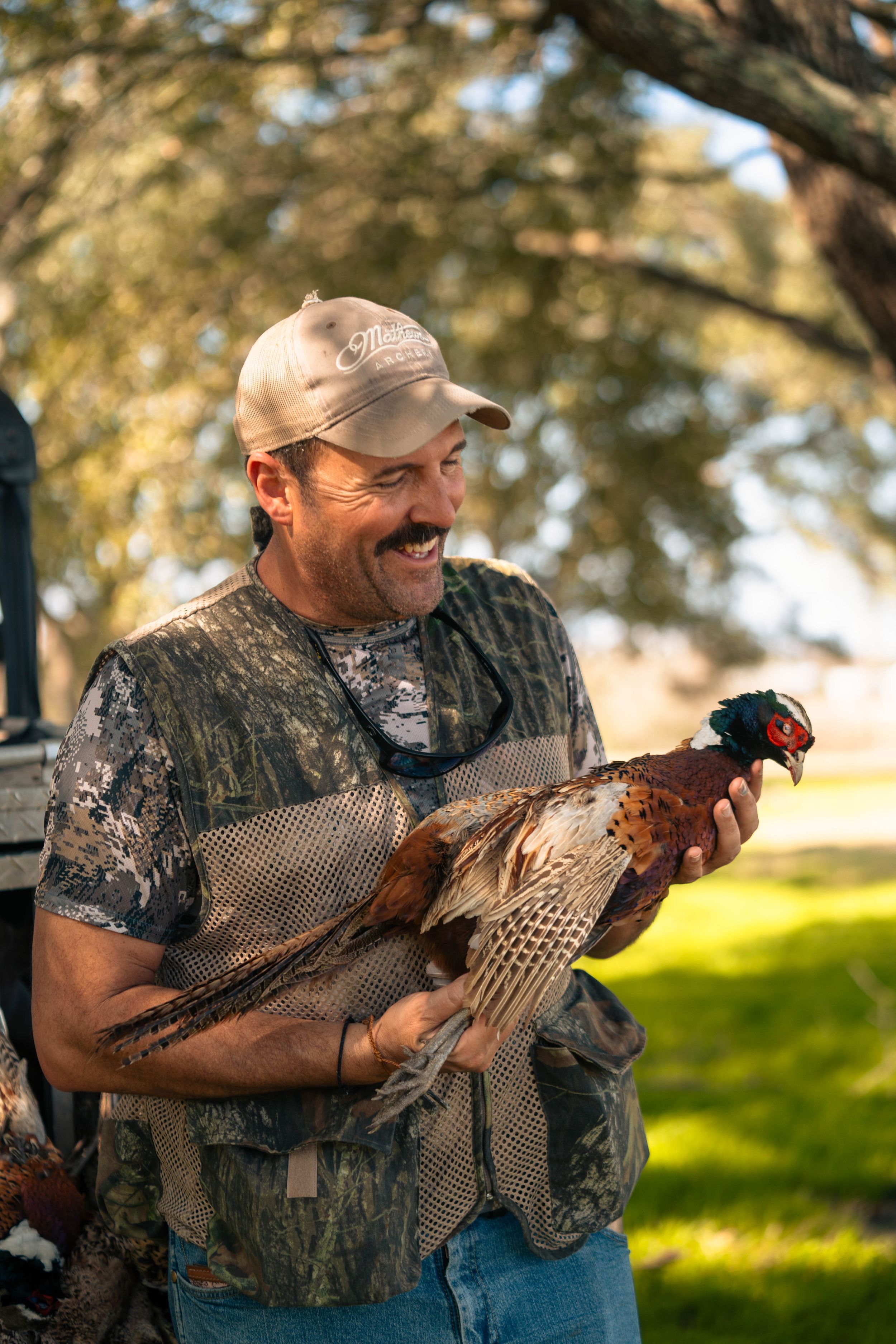 Hunter in camouflage clothing smiles while holding a pheasant in an outdoor setting.