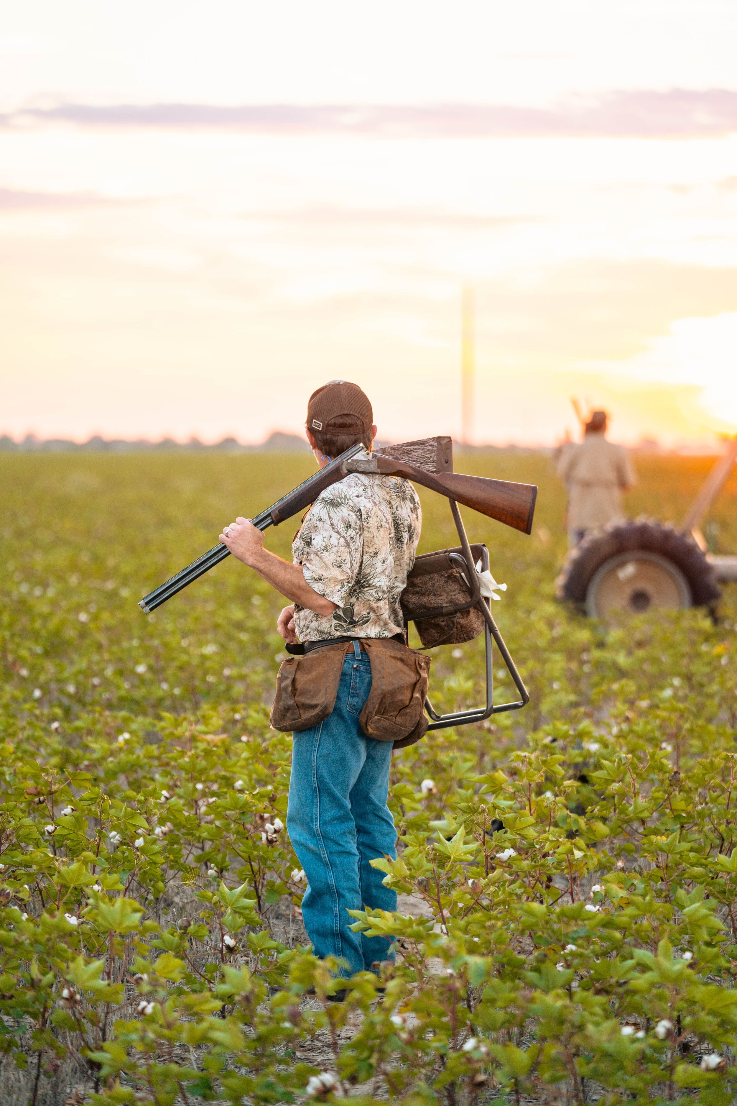 Hunter carrying shotgun walks through field at sunset with tractor in background.
