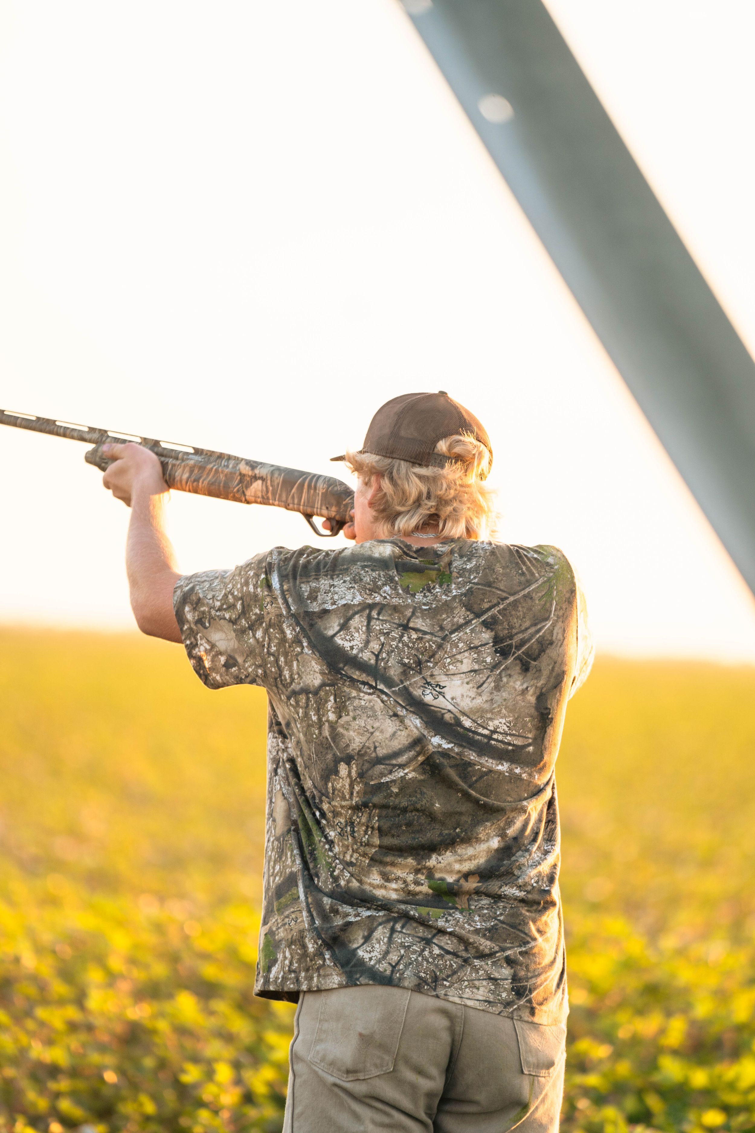 Hunter in camouflage taking aim with shotgun in sunlit field.