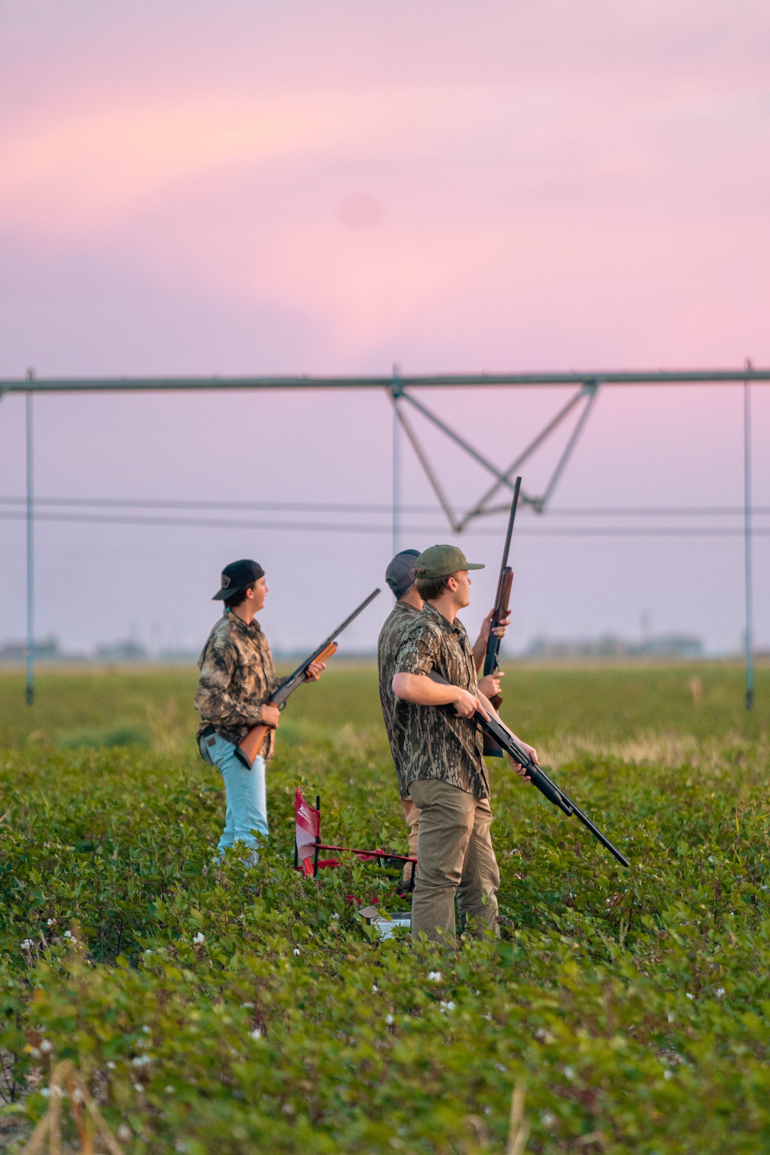 Two hunters in camouflage gear stand in field during pink sunset.
