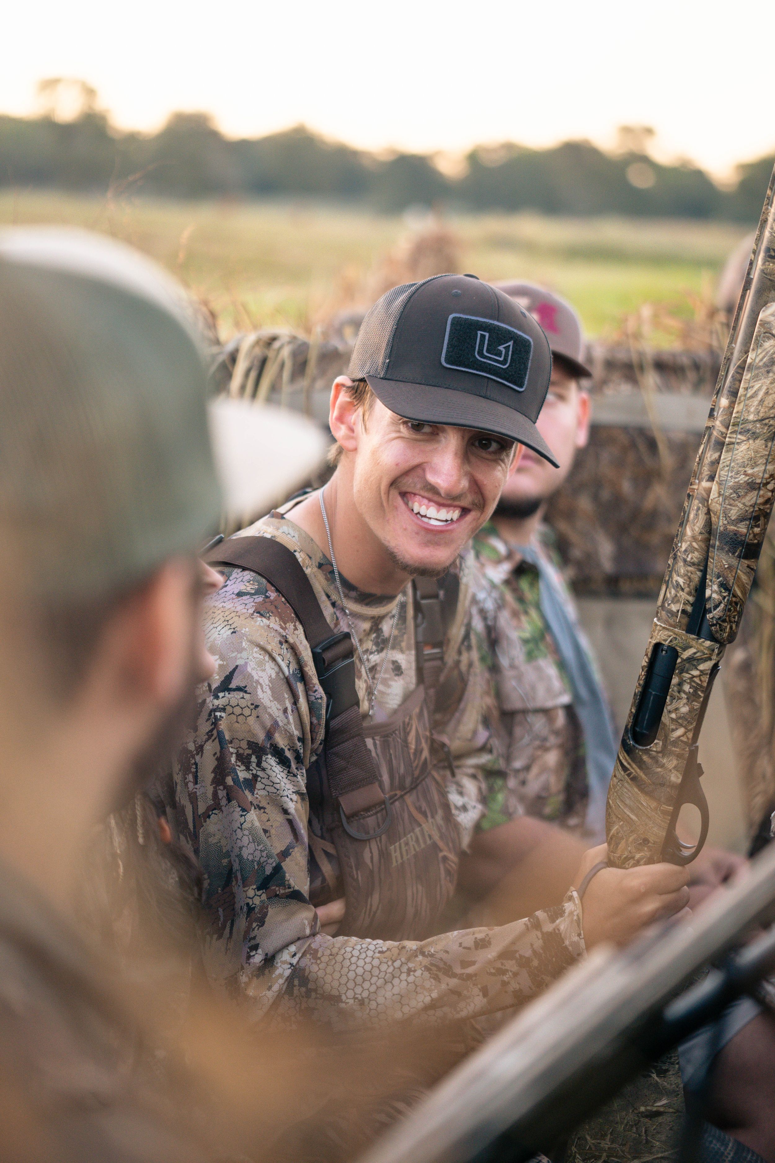 Duck hunter wearing camouflage and baseball cap smiles while in hunting blind.