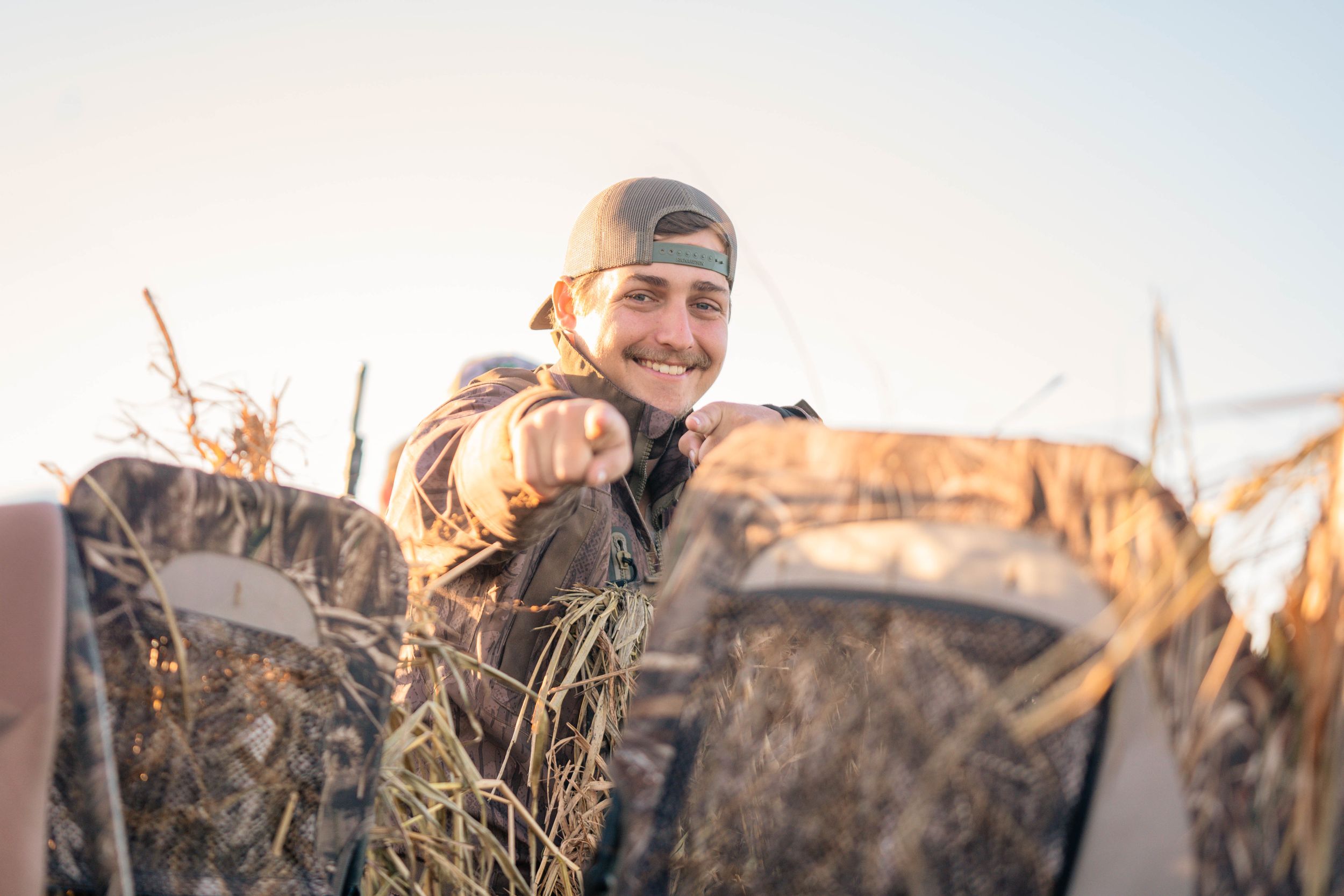 Hunter in camo gear smiles while sitting in duck blind during sunrise.