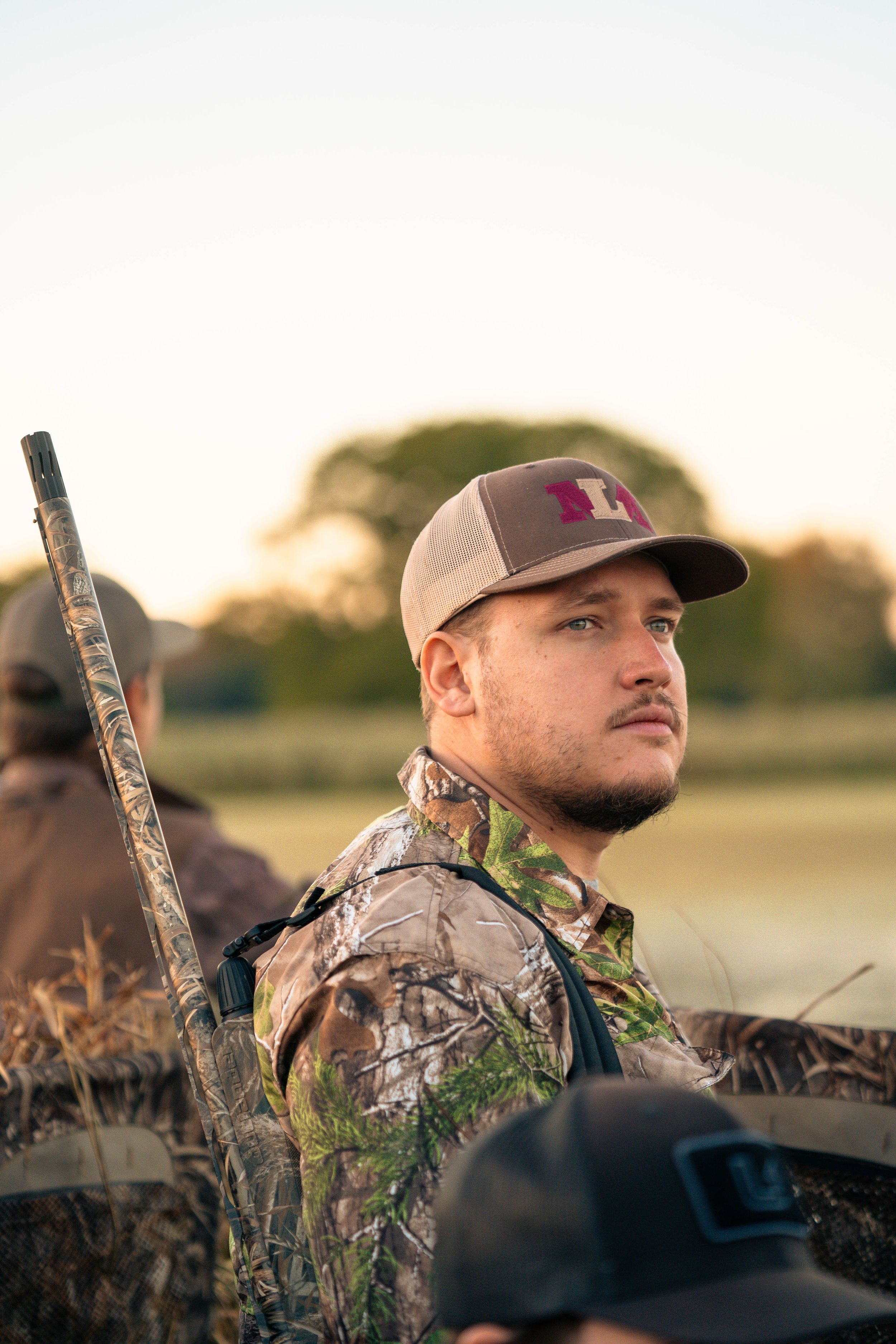 Hunter in camo gear and baseball cap sits in hunting blind during sunrise.