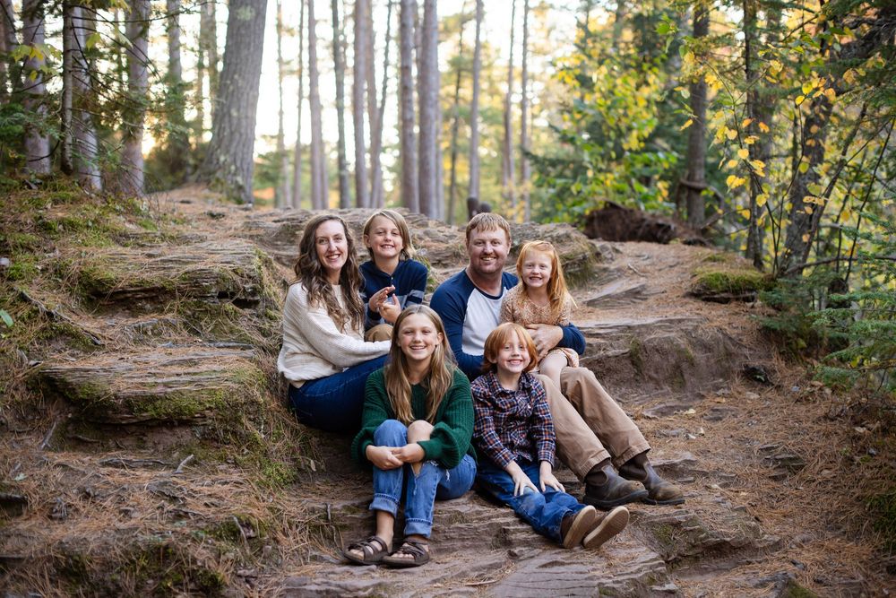 A family sits together on a fallen tree trunk along a wooded hiking trail during an autumn day.