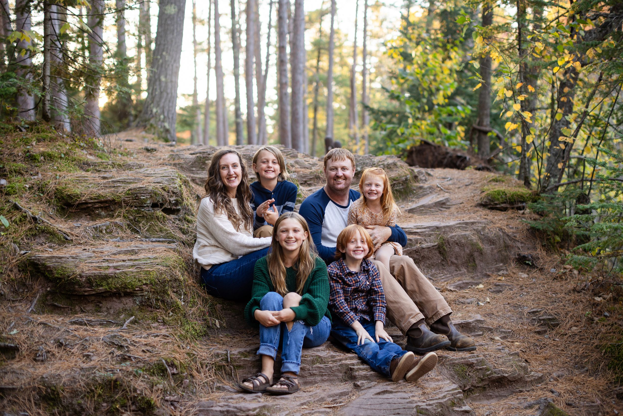 A family sits together on a fallen tree trunk along a wooded hiking trail during an autumn day.