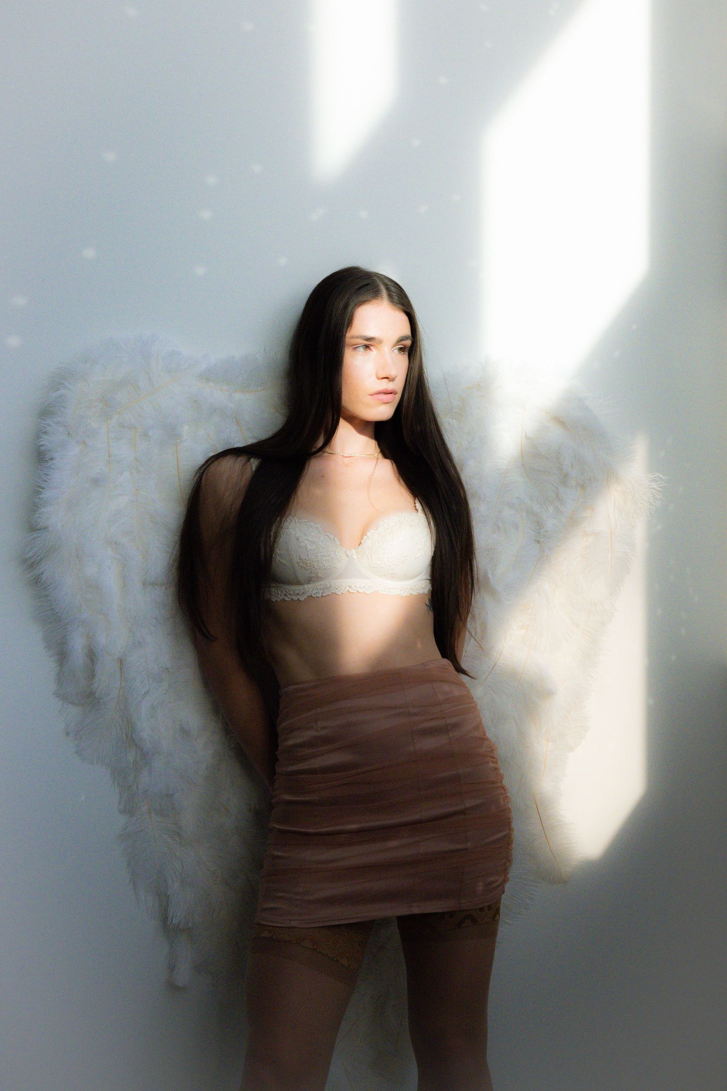 A young woman with long dark hair wears a white bra and sheer skirt with angel wings.