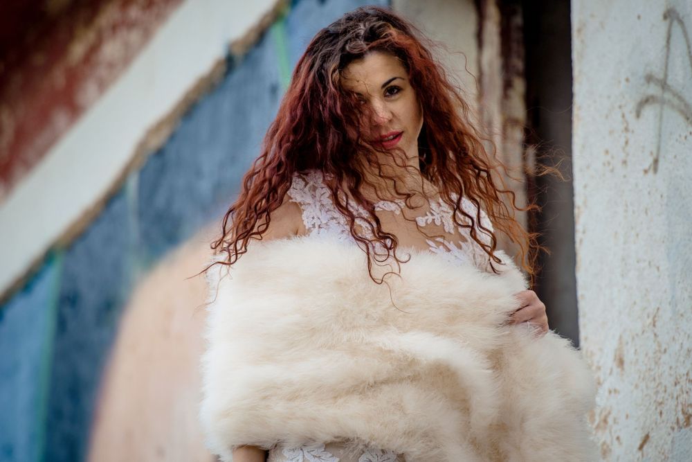 Long curly red hair flowing over a white fuzzy coat while posing against a blue and white textured wall.