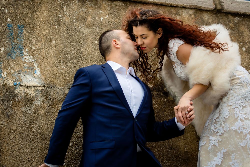 A romantic wedding scene showing a couple in formal attire posing against a weathered wall.