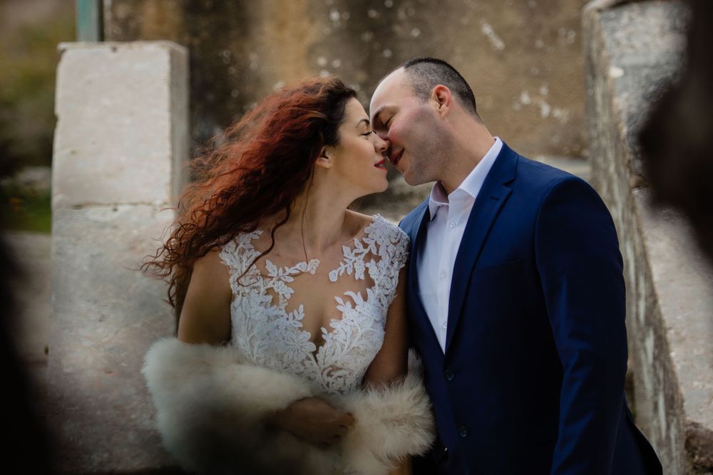 A romantic wedding couple laughs together in front of a stone wall, with the bride wearing a sheer lace gown.