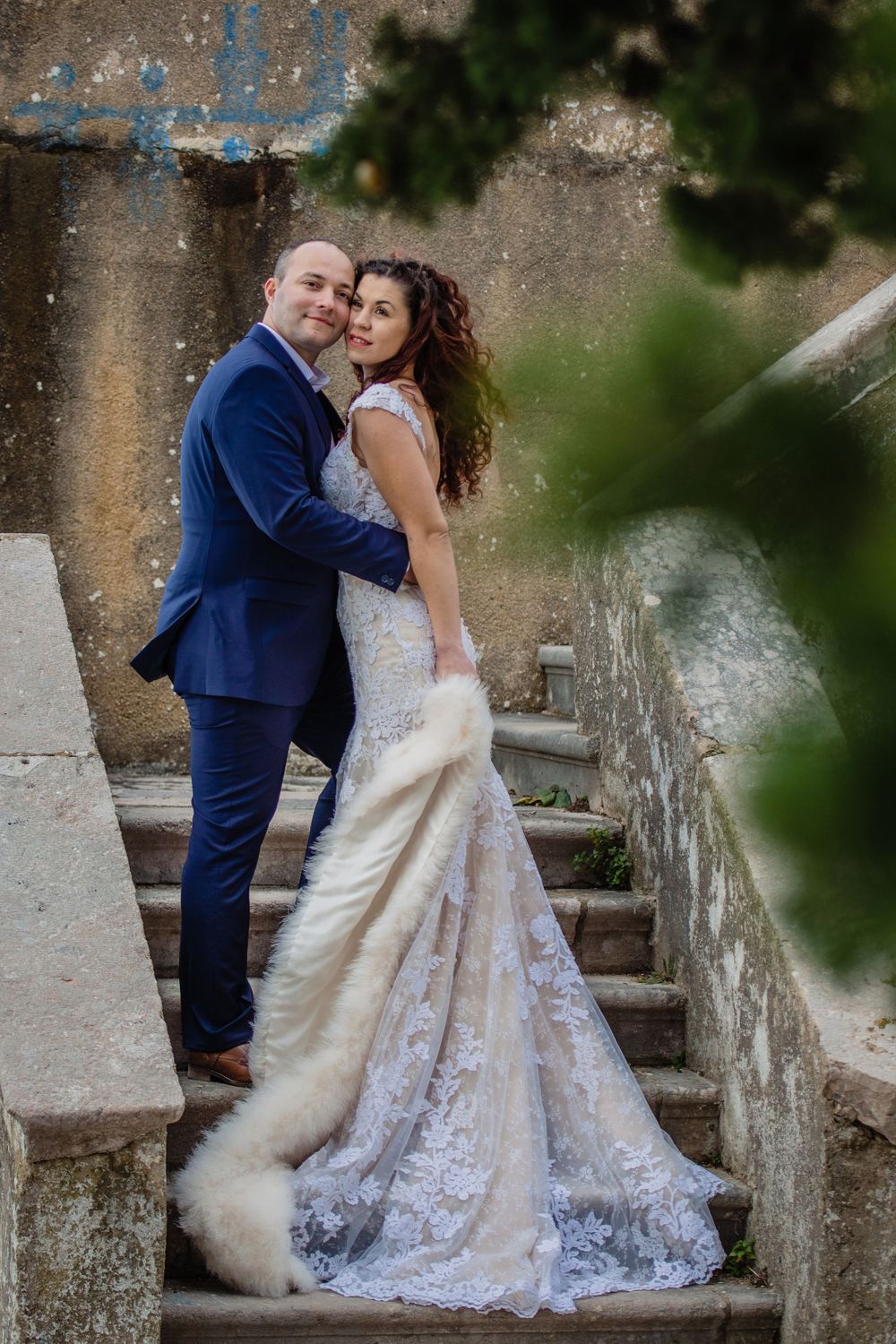 A couple in wedding attire poses on stone steps, the bride wearing a lace dress with long train against an elegant backdrop.